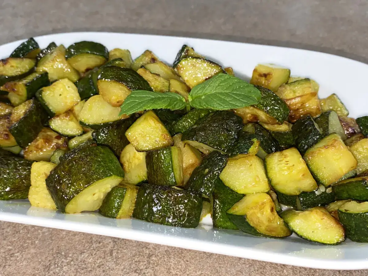 Un piatto di zucchine trifolate, tagliate a cubetti e condite con erbe aromatiche, servite su un piatto bianco.