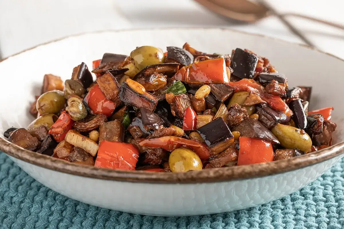 Un piatto invitante di caponata di verdure, con melanzane, peperoni rossi, olive verdi e capperi, condita con un sugo agrodolce.