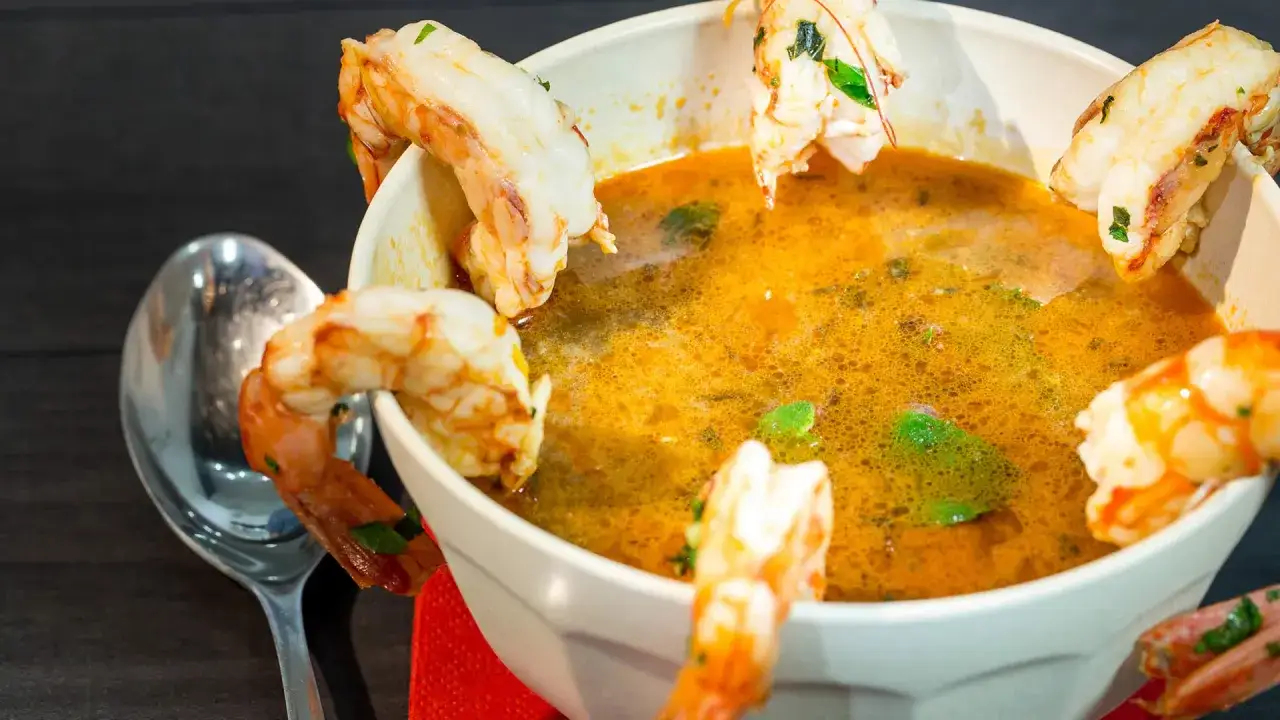 Una ricca zuppa di crostacei, con gamberi succulenti che decorano il bordo del piatto. Un cucchiaio d'argento attende accanto.