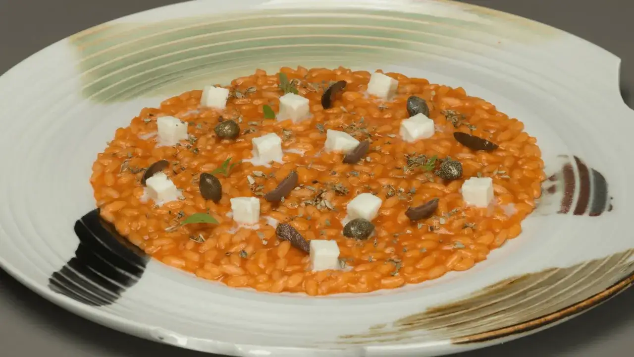 Risotto alla pizzaiola cremoso, guarnito con cubetti di formaggio, olive, capperi e erbe aromatiche su un piatto bianco decorato.