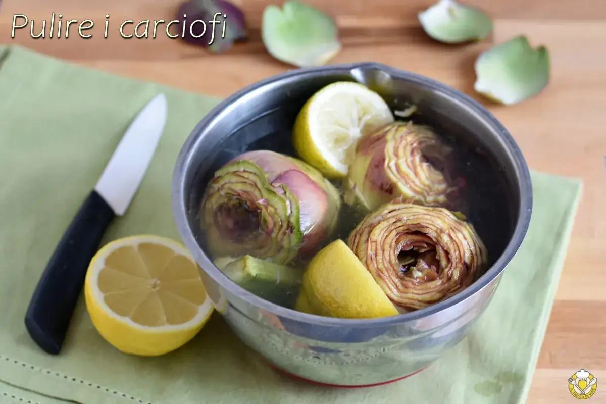 Carciofi puliti e immersi in acqua e limone, pronti per gustose ricette. Un limone tagliato a metà e un coltello completano la scena.