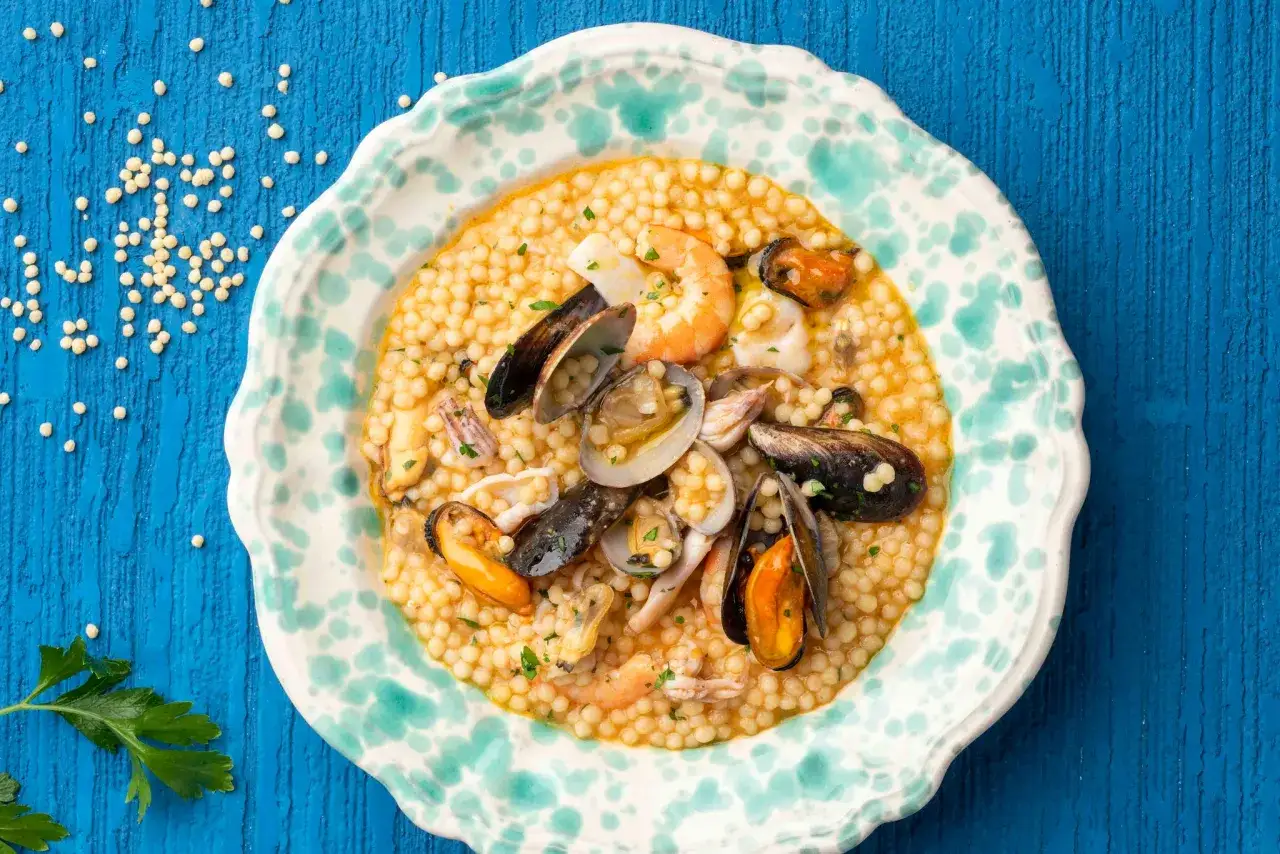 Un piatto di fregola sarda con frutti di mare, cozze, vongole e gamberi, guarnito con prezzemolo fresco.