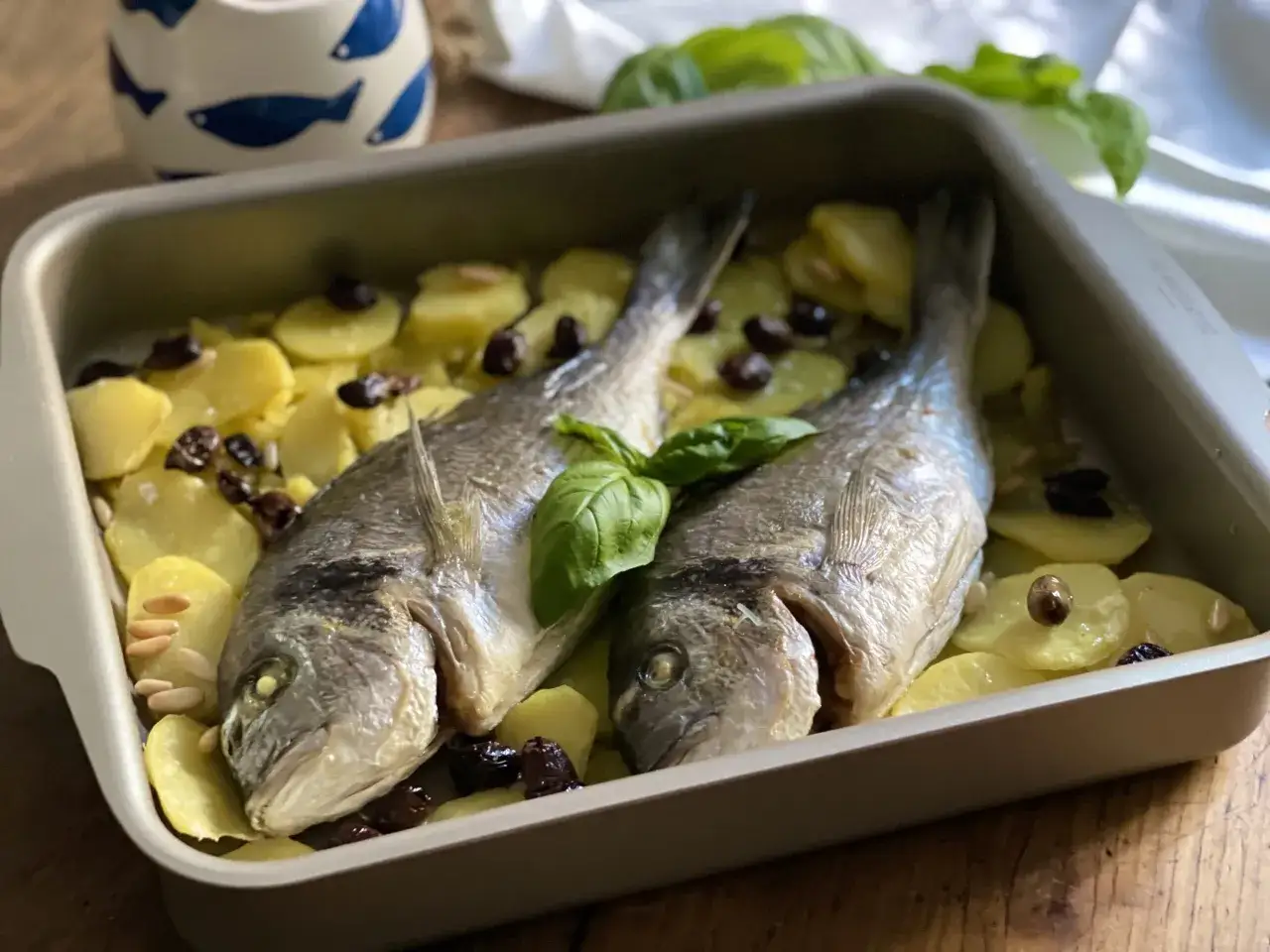 Orata alla ligure appena sfornata, con patate, olive e pinoli, guarnita con basilico fresco.