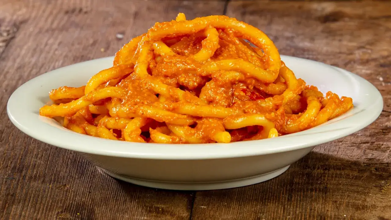 Pici al sugo all'aglione, un piatto tradizionale toscano servito in una ciotola bianca su un tavolo di legno rustico.