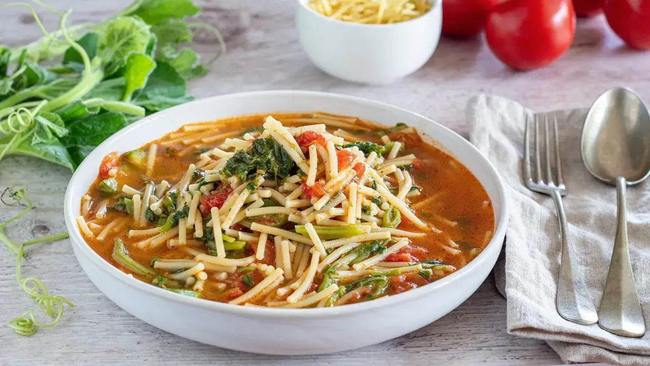 A white bowl of pasta with tenerumi, tomatoes, and a rich broth, garnished with fresh greens.