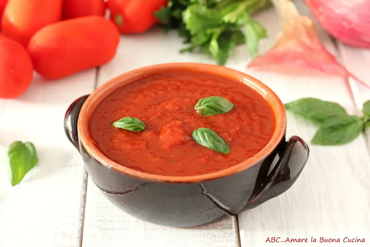 Pomarola ricetta: salsa di pomodoro fatta in casa in ciotola di terracotta, guarnita con basilico fresco. Ingredienti freschi sullo sfondo.
