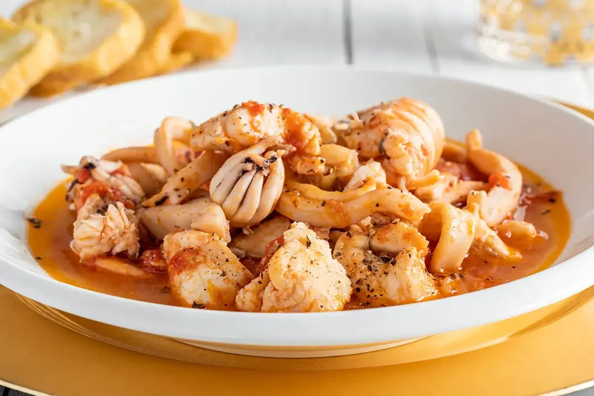 Un invitante piatto di zuppa di pesce surgelato, con calamari, gamberi e pezzi di pesce bianco in un sugo rosso, servito con pane tostato.