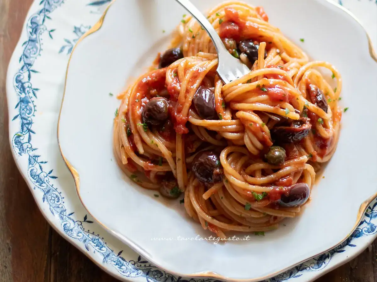 Spaghetti al sugo alla puttanesca, con pomodoro, olive nere e capperi, serviti su un piatto bianco con decorazioni blu.