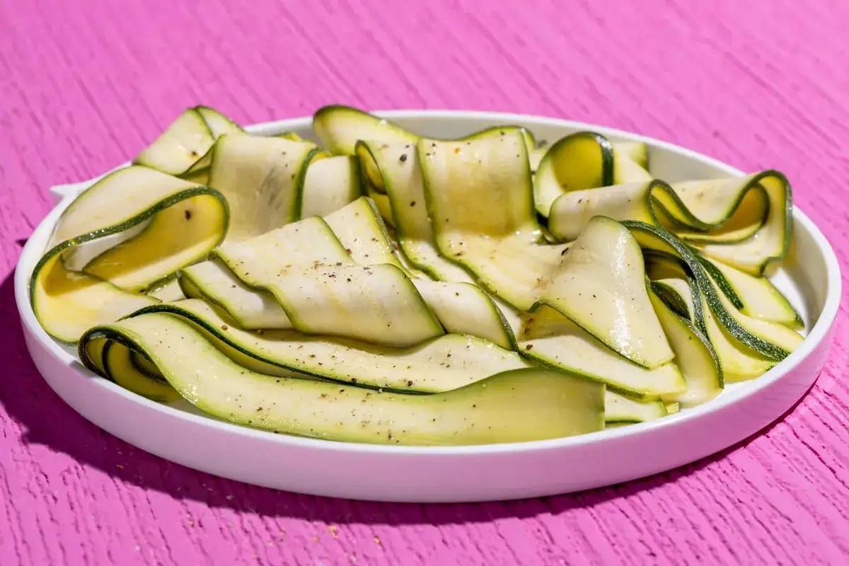 Fettine sottili di zucchine marinate, condite con pepe nero, disposte a spirale in un piatto bianco su un tavolo rosa acceso.