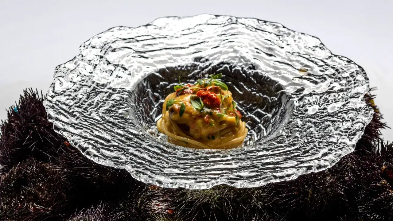 Un piatto di spaghetti conditi con una crema delicata e pomodorini, servito su un letto di ricci di mare freschi, il tutto incorniciato da un'elegante ciotola di vetro.