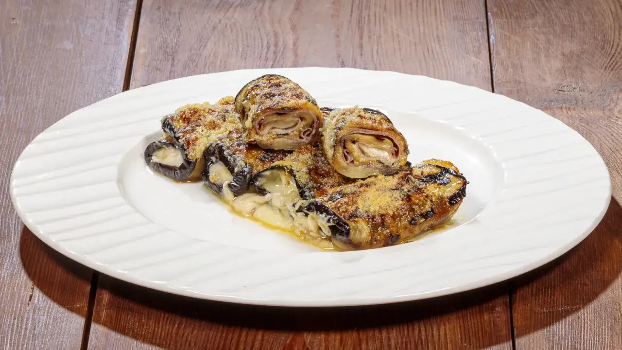 Involtini di melanzane grigliate ripieni di formaggio e prosciutto, spolverati di parmigiano, su un piatto bianco.