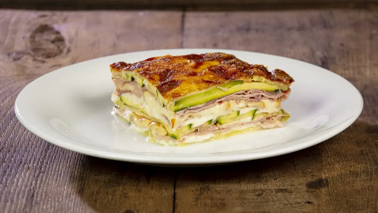 Una fetta di parmigiana di zucchine, stratificata con prosciutto e formaggio filante, dorata al forno su un piatto bianco.
