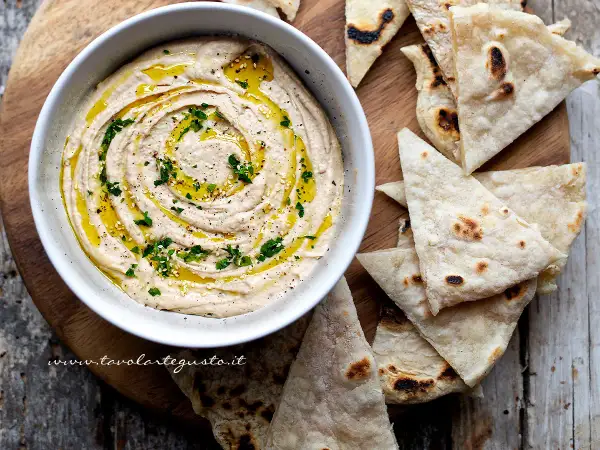 Delizioso hummus di fagioli cremoso, condito con olio d'oliva, prezzemolo e semi di sesamo, servito con pane pita tostato.