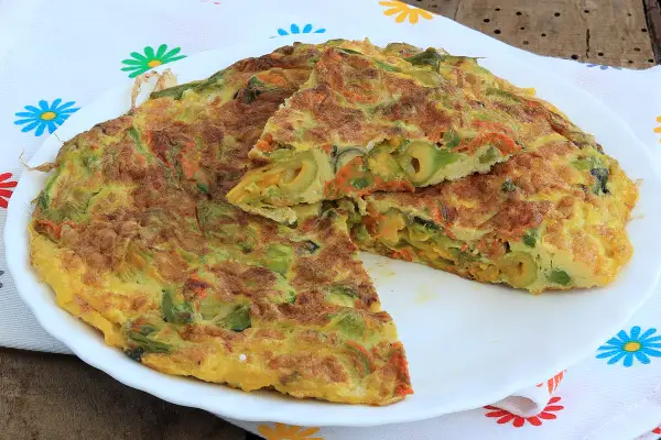 Frittata con fiori di zucca, carote e olive, tagliata a spicchi su un piatto bianco, con un tovagliolo colorato sullo sfondo.