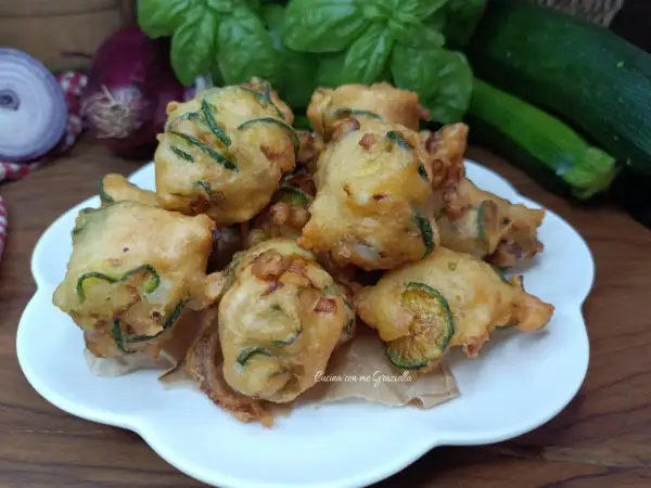 Frittelle di zucchine dorate e croccanti, pronte per essere gustate. Sullo sfondo, ingredienti freschi come basilico e cipolle rosse.