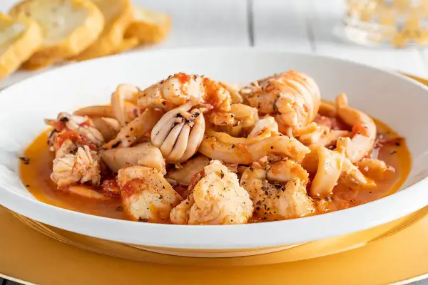 Un invitante piatto di zuppa di pesce surgelato, con calamari, gamberi e pezzi di pesce bianco in un sugo rosso, servito con pane tostato.