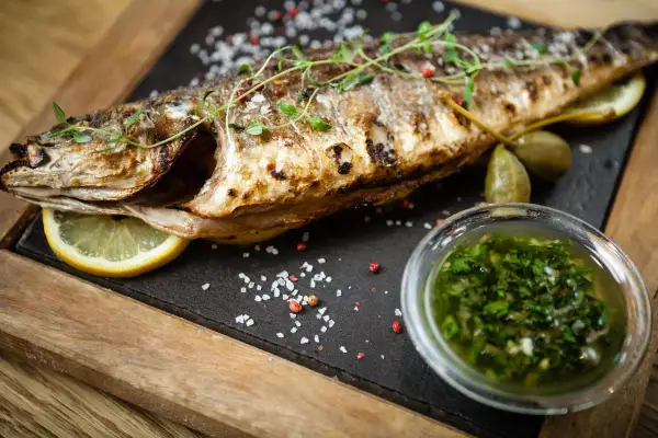Un succulento branzino al forno, guarnito con erbe aromatiche, limone e capperi, accompagnato da una salsa verde.