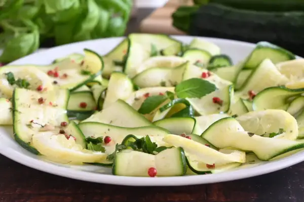 Fette di zucchine marinate, condite con pepe rosa e erbe aromatiche, su un piatto bianco.
