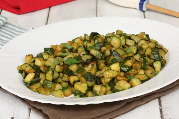 Zucchine in padella, tagliate a cubetti e condite con erbe aromatiche fresche, servite su un piatto bianco.