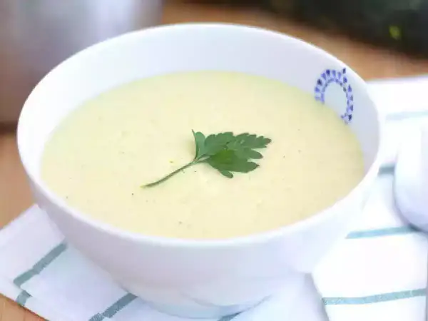 Una cremosa vellutata di zucchine, guarnita con un rametto di prezzemolo fresco, servita in una ciotola bianca con un bordo decorato.