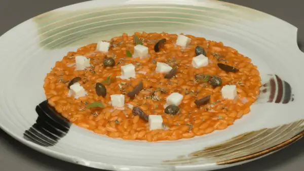 Risotto alla pizzaiola cremoso, guarnito con cubetti di formaggio, olive, capperi e erbe aromatiche su un piatto bianco decorato.
