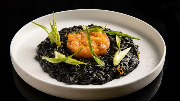 Un piatto di risotto al nero di seppia, sormontato da tartare di salmone e guarnito con erbe fresche e scorza d'arancia.