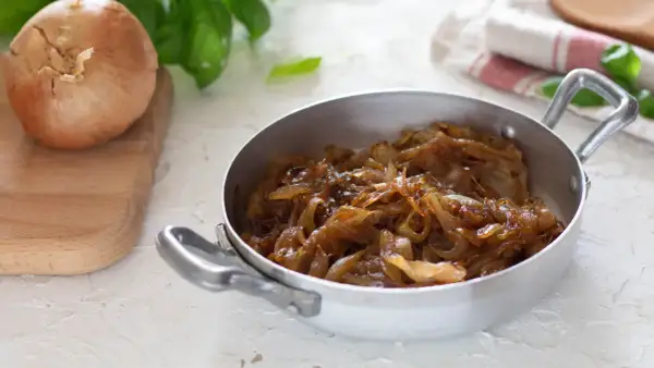 Cipolle caramellate in padella, pronte per insaporire un piatto. Accanto, una cipolla cruda su un tagliere.