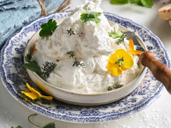 Burro montato soffice e arioso, guarnito con erbe aromatiche e fiori gialli, servito in una ciotola decorata.