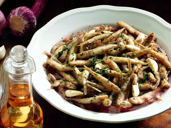 Un saporito carpione piemontese in un piatto bianco, condito con aglio e prezzemolo, accanto a una bottiglia d'olio e aglio viola.