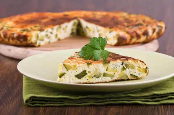 Una fetta di frittata di zucchine, soffice e dorata, guarnita con prezzemolo fresco, su un piatto chiaro.
