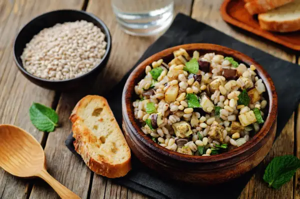 Insalata di farro con verdure miste, pinoli e menta, servita in una ciotola di legno. Accanto, pane tostato e una ciotola di farro crudo.