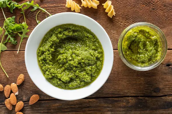 Pesto di rucola e mandorle in una ciotola bianca, accanto a un vasetto pieno, con mandorle e rucola fresca sparse sul tavolo di legno.