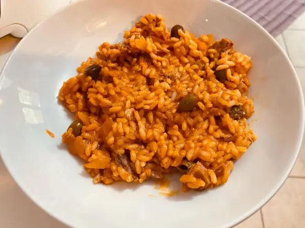 Un invitante risotto al pomodoro, ricco e saporito, con pezzetti di carne e olive nere, servito in una ciotola bianca.