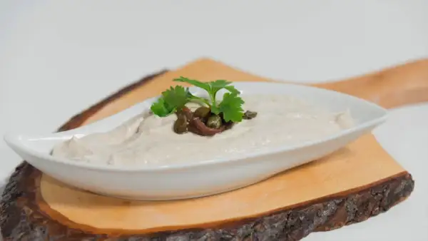Salsa tonnata cremosa, guarnita con acciughe, capperi e prezzemolo, servita in una ciotola bianca su un tagliere di legno.