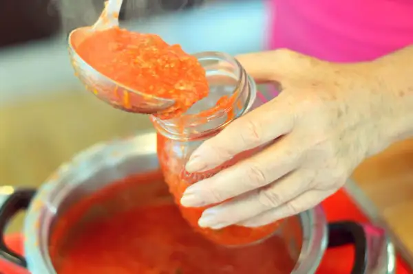 Mani che versano pomarola ricetta in un barattolo di vetro. Il sugo fumante viene preso da una pentola con un mestolo.